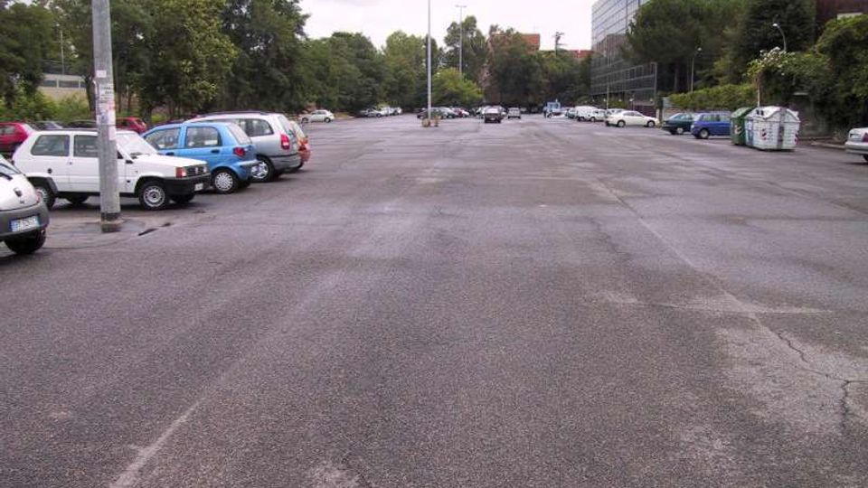 Ampio parcheggio asfaltato all’aperto con auto parcheggiate lungo i lati e corsia centrale libera. Sullo sfondo ci sono alberi e, a destra, un edificio moderno con facciata in vetro e alcune aree verdi ai margini.