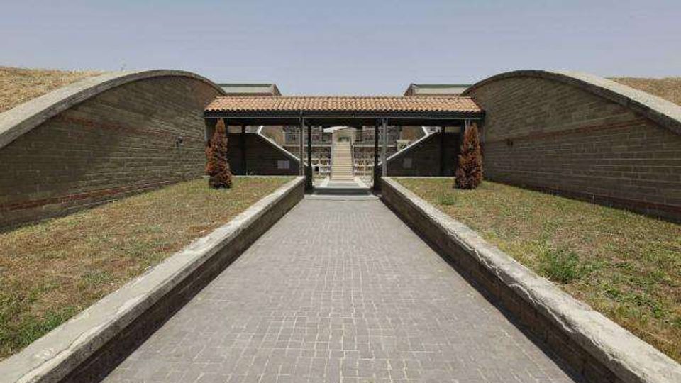 Ingresso a un complesso architettonico con un lungo viale pavimentato in mattoni tra basse aiuole erbose, affiancato da due grandi muri curvi in mattoni. In fondo si vede un portico con tetto in tegole sostenuto da pilastri e l’accesso a un edificio interno.