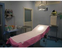 Sala medica/ambulatorio con lettino da visita coperto da lenzuolo bianco e telo rosa, illuminato da una lampada operatoria a soffitto. Sulle pareti sono presenti monitor e apparecchiature mediche, con carrelli e un tavolo con strumenti e materiali.