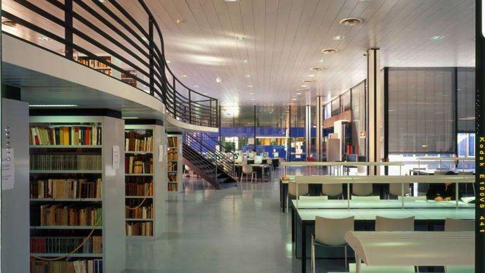 Interno di una biblioteca moderna con scaffali di libri, tavoli di studio con lampade e sedie disposti in file. Si vedono un soppalco con ringhiera metallica e una scala che conduce al livello superiore, sotto un soffitto alto con illuminazione integrata.