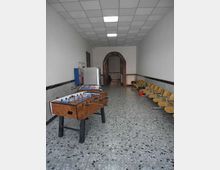 Sala ricreativa rettangolare con pavimento in graniglia e soffitto a pannelli, con due biliardini al centro e una fila di sedie gialle lungo la parete destra. In fondo si vede un arco in mattoni che conduce a un’altra stanza, con pareti bianche rivestite a mezza altezza.