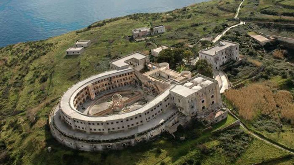 Veduta aerea di un grande complesso edilizio semicircolare in muratura, con cortile interno e ali laterali, situato su una collina verde vicino alla costa. Intorno si vedono strade sterrate, campi terrazzati e pochi edifici isolati, con il mare sullo sfondo.