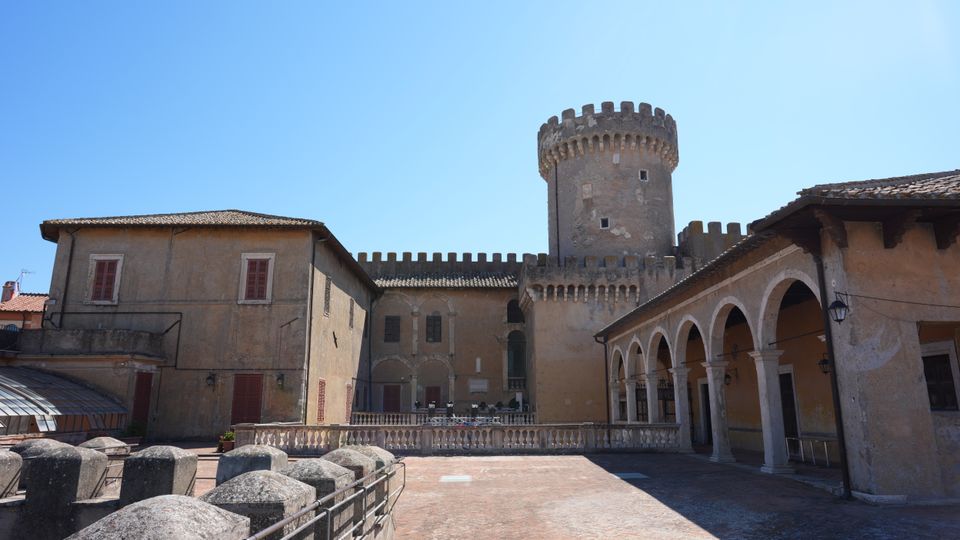 Cortile interno di un castello in muratura con torre cilindrica merlata e mura fortificate sullo sfondo. A destra corre un porticato con archi e colonne, mentre al centro si apre un’ampia terrazza pavimentata con balaustra in pietra.