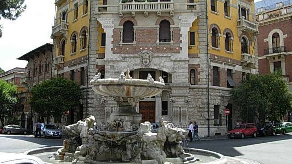 Fontana monumentale in pietra al centro di una rotonda stradale, con vasca circolare e sculture decorative alla base. Sullo sfondo si vede un grande edificio storico a più piani con facciata gialla e dettagli in pietra, archi e balconi, affacciato su un incrocio urbano.