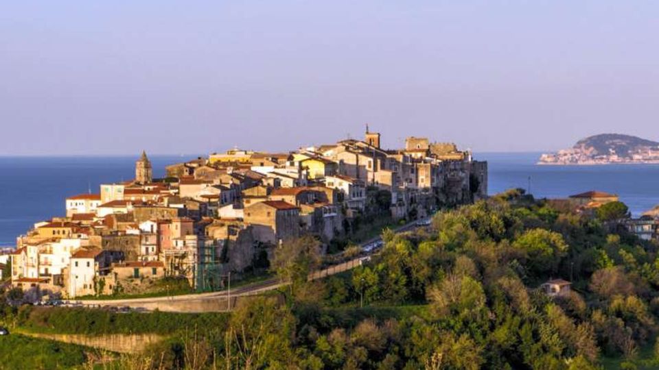 Veduta panoramica di un borgo medievale su un crinale, con case in pietra addossate tra loro e una torre campanaria, circondato da colline verdi. Sullo sfondo si vede il mare con una costa lontana sotto un cielo sereno.