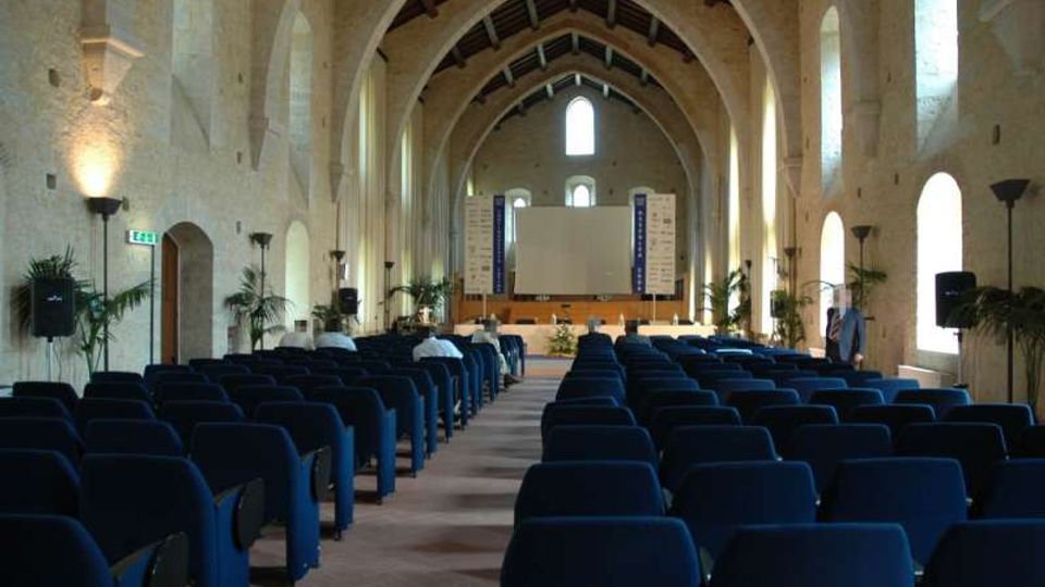 Interno di una grande sala conferenze ricavata in un edificio in pietra con alte arcate e soffitto a capriate, illuminata da finestre ad arco sui lati. File di poltrone blu sono orientate verso un palco con grande schermo di proiezione e pannelli informativi sullo sfondo.
