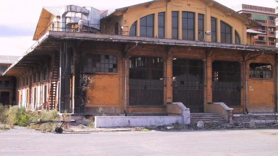 Edificio industriale in mattoni con grande tetto a capanna e ampie finestre ad arco, con portoni metallici e una veranda coperta su pilastri lungo la facciata. Area esterna asfaltata e spoglia, con segni di degrado e strutture metalliche visibili sul lato.