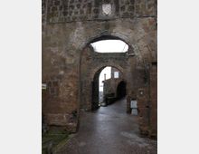 Arco in pietra di una porta medievale con stemma scolpito, che attraversa una spessa muratura in blocchi. Sotto l’arco si vede un secondo passaggio ad arco e una stretta strada lastricata in leggera discesa tra edifici in pietra.