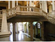 Interno di un edificio storico con grande scala in pietra e ballatoio con balaustra, sostenuti da colonne. Al piano terra si vedono archi e porte vetrate ad arco che si affacciano su un atrio luminoso.