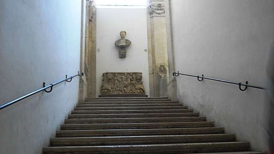 Scalinata interna in pietra con corrimano laterali, che conduce a un pianerottolo illuminato da una finestra ad arco sotto una volta. Sulla parete di fondo sono visibili un busto scultoreo e un bassorilievo decorativo tra elementi architettonici in pietra.