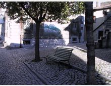Piccola piazzetta con pavimentazione in ciottoli, una panchina metallica e alberi che creano ombra. Sullo sfondo si vedono facciate di edifici in pietra con finestre grigliate, una scala esterna e un murale dipinto sul muro.