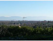 Veduta panoramica di una campagna con vegetazione bassa in primo piano e alcune case sparse e antenne sullo sfondo. All’orizzonte si distinguono colline e una catena montuosa in lontananza sotto un cielo sereno.