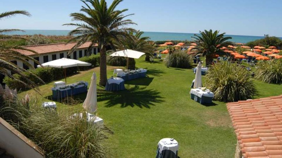 Giardino di un resort sul mare con ampio prato verde, palme e tavoli apparecchiati sotto grandi ombrelloni bianchi. Sullo sfondo si vede la spiaggia con numerosi ombrelloni arancioni e edifici a un piano con tetti in tegole rosse.