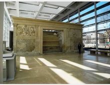 Interno di un museo o atrio moderno con ampie vetrate a tutta altezza e soffitto a griglia, che ospita una grande struttura in pietra scolpita con bassorilievi e un’apertura centrale. Pavimento lucido in pietra con fasci di luce e ombre proiettate dalle finestre.