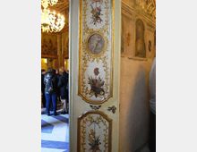 Porta interna decorata in stile barocco, dipinta con medaglioni, ghirlande dorate e motivi araldici, che si apre su una sala storica con lampadario e pareti affrescate. Sullo sfondo si intravedono pavimento a scacchi e ambienti riccamente ornati.