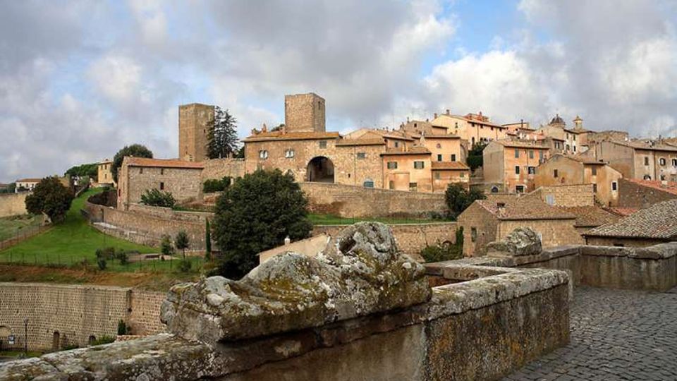 Vista panoramica di un borgo medievale su una collina, con case in pietra dai tetti in tegole, mura fortificate e una torre quadrata. In primo piano si vede un parapetto in pietra e un camminamento lastricato, con campi verdi ai piedi delle mura sotto un cielo nuvoloso.