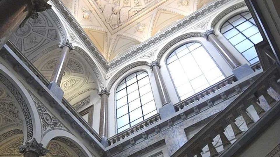 Interno di un edificio storico con grande vano scala in pietra, balaustre e colonne decorate, visto dal basso verso una cupola a cassettoni con lucernario centrale. Alte finestre ad arco illuminano le pareti ornate con stucchi e motivi classici.