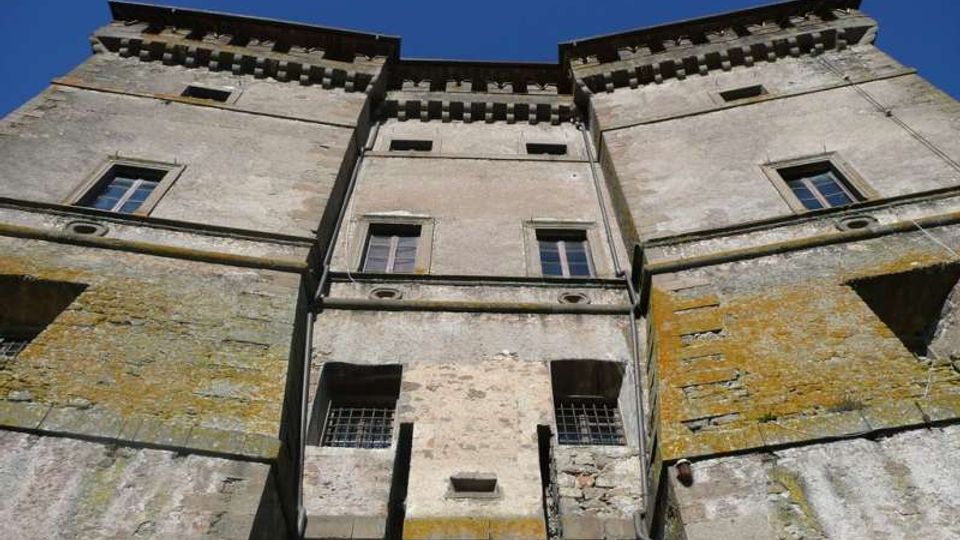 Vista dal basso della facciata di un castello in pietra con due torri laterali e corpo centrale, finestre rettangolari e merli sotto la gronda. Le pareti mostrano intonaco e pietra invecchiati con tracce di muschio, sullo sfondo di un cielo azzurro.