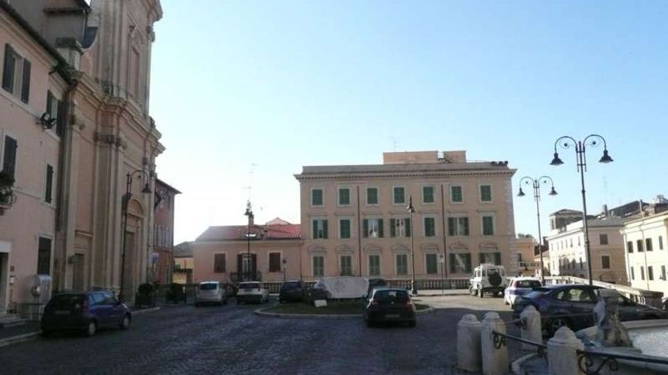 Piazza lastricata in pietra con auto parcheggiate, circondata da edifici storici: a sinistra una chiesa con facciata alta e decorata, di fronte un palazzo rettangolare con persiane verdi. Lampioni in ferro e una piccola area con balaustra in primo piano definiscono il bordo della piazza sotto un cielo sereno.