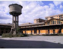 Grande cortile industriale con un alto serbatoio d’acqua cilindrico su pilastri in cemento e un edificio basso a pianta semicircolare con facciata arancione e finestre ad arco. Sullo sfondo si vedono palazzi residenziali e cielo parzialmente nuvoloso.