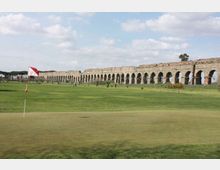Campo da golf con bandierina sul green in primo piano, circondato da ampie aree erbose. Sullo sfondo si estende un lungo acquedotto in pietra con una serie di arcate sotto un cielo parzialmente nuvoloso.