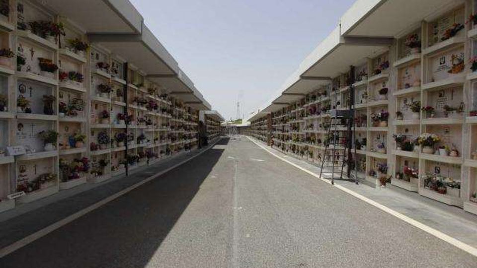 Viale centrale asfaltato di un cimitero, fiancheggiato su entrambi i lati da lunghe strutture a più livelli con loculi e lapidi. Le pareti dei colombari sono decorate con fiori e targhe, con prospettiva che converge verso l’orizzonte sotto un cielo sereno.