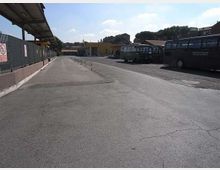 Ampio piazzale asfaltato di un deposito autobus, con pensilina e recinzione sul lato sinistro e vari autobus parcheggiati lungo il lato destro. Sullo sfondo si vedono edifici bassi e una collina alberata sotto un cielo sereno.