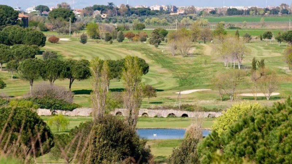 Ampio campo da golf con fairway ondulati, alberi sparsi e alcune aree di sabbia, con un piccolo laghetto in primo piano. Sullo sfondo si vedono campi agricoli e un’area urbana lontana all’orizzonte.