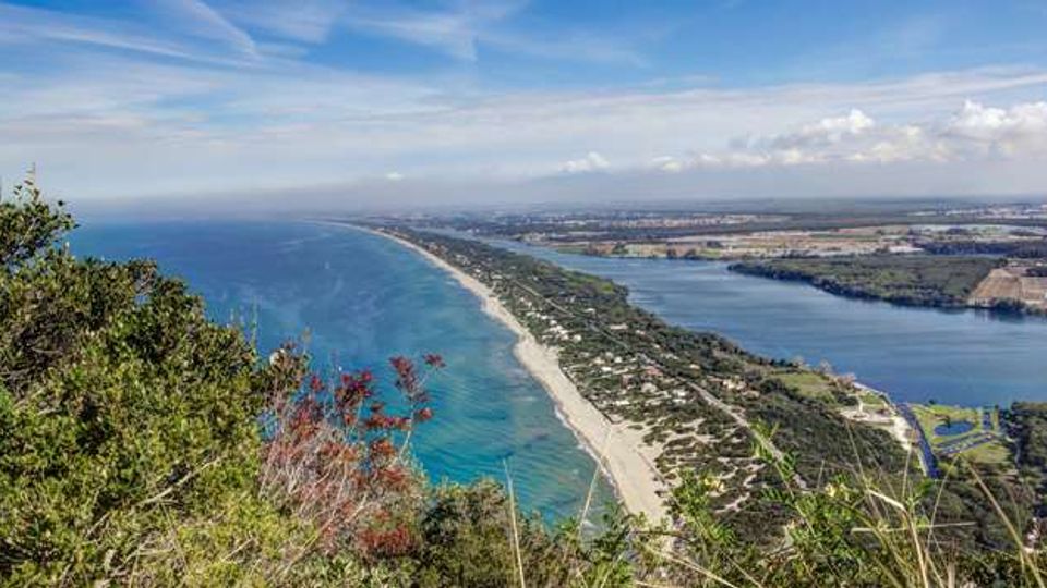Veduta panoramica dall’alto di un lungo litorale sabbioso con mare turchese, affiancato da una fascia di vegetazione costiera e strade. Sul lato opposto si estende una grande laguna o lago costiero, con pianure e campi visibili all’orizzonte sotto un cielo sereno.