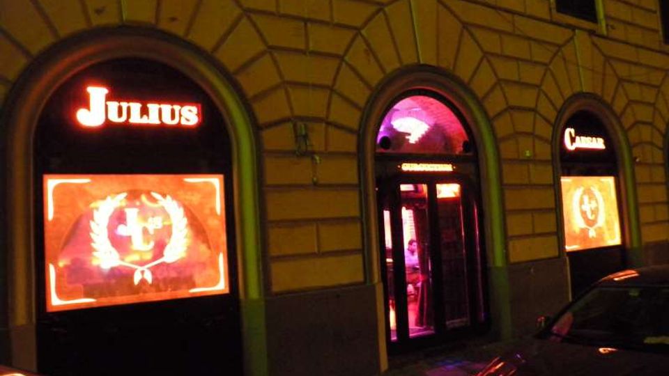 Facciata notturna di un edificio urbano con grandi finestre ad arco e ingresso centrale illuminato da luci rosse e viola. Sulle vetrine laterali sono visibili insegne luminose e grafiche, con auto parcheggiate lungo la strada.