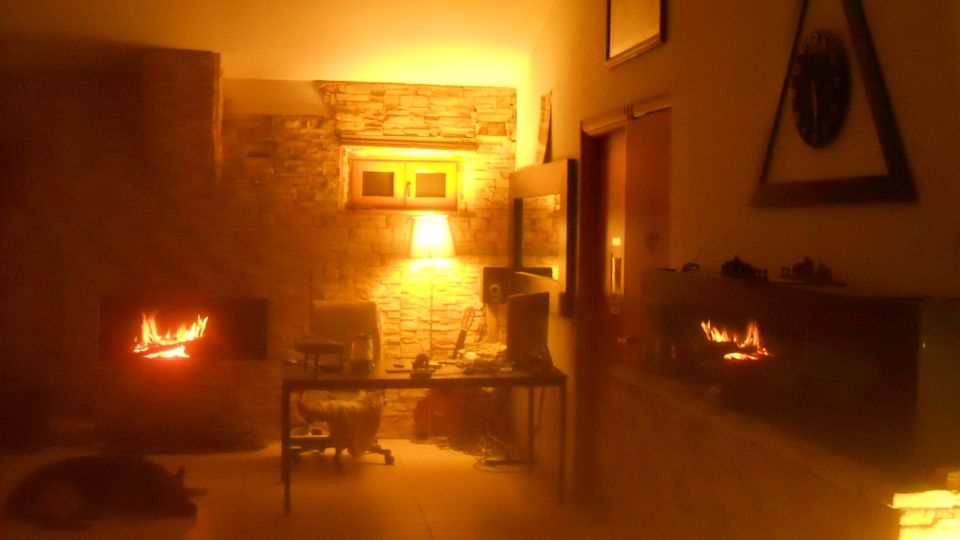 Ampio soggiorno con pareti in pietra e due camini accesi, con illuminazione calda e soffusa. Al centro c’è un tavolo con sedie e oggetti, e sul fondo si vede una piccola finestra incassata nel muro.