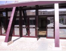 Ingresso di un edificio moderno con grandi vetrate e telai metallici inclinati color bordeaux, con porte in vetro che si aprono su un atrio interno. Davanti si vede una pavimentazione in pietra e, sullo sfondo, un cortile con altri edifici.