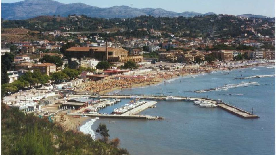 Veduta panoramica di una cittadina costiera con lunga spiaggia attrezzata e un porto turistico con moli e barche. Sullo sfondo si vedono edifici e colline verdeggianti che salgono verso montagne lontane.