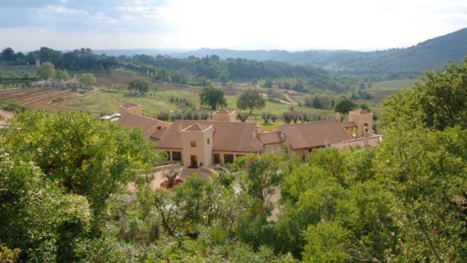Vista panoramica di un complesso di edifici rurali con tetti in tegole e piccoli campanili, circondato da vegetazione. Sullo sfondo si estendono campi coltivati, filari di alberi e colline verdi sotto un cielo parzialmente nuvoloso.