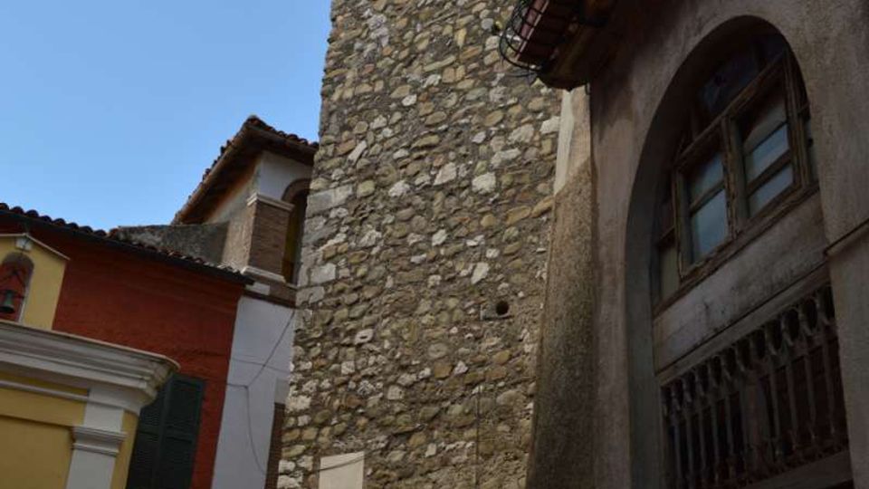 Torre alta in pietra grezza con piccole aperture in alto, affacciata su un vicolo stretto tra edifici colorati. A destra si vede una facciata con grande portone ad arco e balcone con fioriere sotto il tetto.