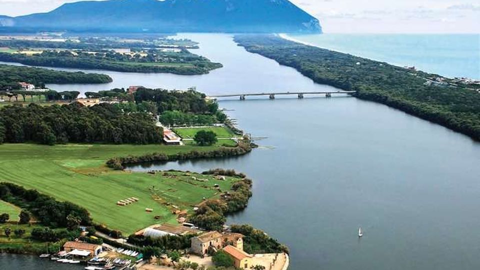 Veduta aerea di un ampio fiume o canale che attraversa una pianura costiera con campi verdi e boschi, collegato da un ponte centrale e diretto verso il mare. In primo piano si vede una piccola penisola con edifici e un molo, mentre sullo sfondo si staglia una grande montagna lungo la costa.