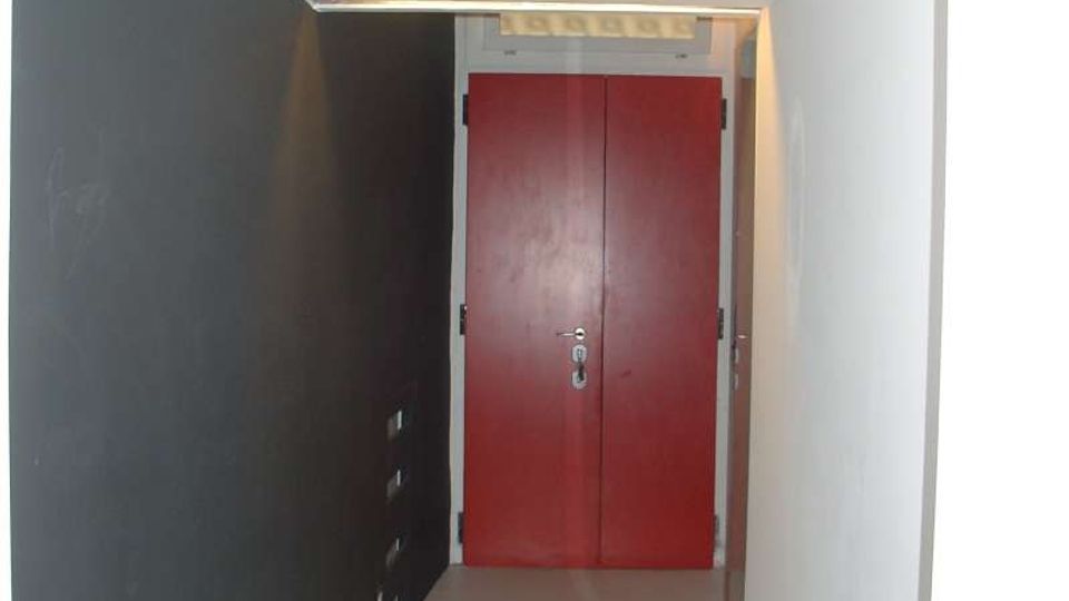 Corridoio interno stretto con pavimento chiaro, parete sinistra scura e parete destra bianca, che conduce a una porta doppia rossa in fondo. Soffitto basso con piccola griglia o vano di aerazione sopra la porta.