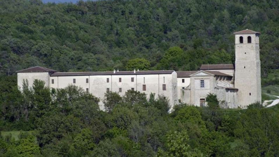 Grande complesso monastico in pietra chiara con edifici allungati e una torre campanaria quadrata, situato ai margini di una collina boscosa. La struttura è circondata da vegetazione fitta e si staglia sullo sfondo di un pendio verde.