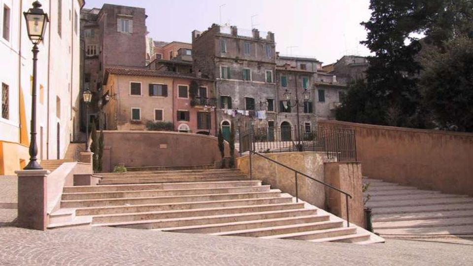Scalinata in pietra che collega una piazzetta pavimentata in sampietrini a una terrazza con muretti e ringhiere. Sullo sfondo si vedono edifici storici con facciate usurate e finestre, lampioni in ferro e alberi al lato destro.