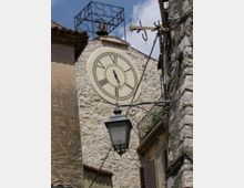 Facciata in pietra di un edificio storico con grande orologio rotondo a numeri romani e una campana in una struttura in ferro sopra il tetto. In primo piano si vede un lampione sospeso e, a destra, un balcone con ringhiera e cavi lungo il muro di una stretta via tra edifici.
