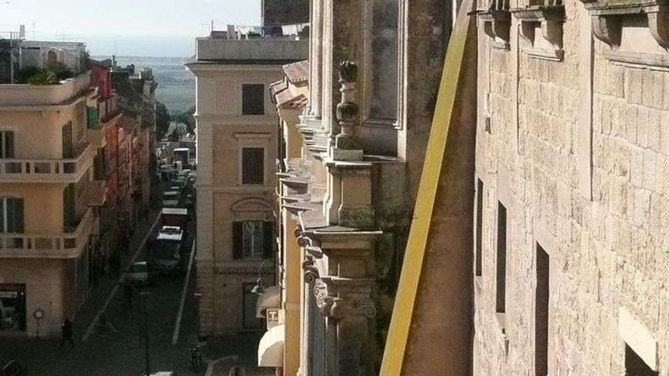 Vista dall’alto di una strada urbana in discesa affiancata da edifici storici in pietra e facciate ornate con balconi e cornici. In fondo si intravede l’orizzonte sul mare, con auto parcheggiate lungo i lati della via.