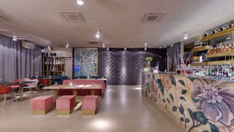 Interno di un lounge bar moderno con bancone sulla destra decorato con grandi motivi floreali e bottiglie esposte su mensole illuminate. A sinistra ci sono tavolini con panche imbottite e sedute, mentre sul fondo una parete scura con texture ondulata e tende grigie incornicia l’ambiente.