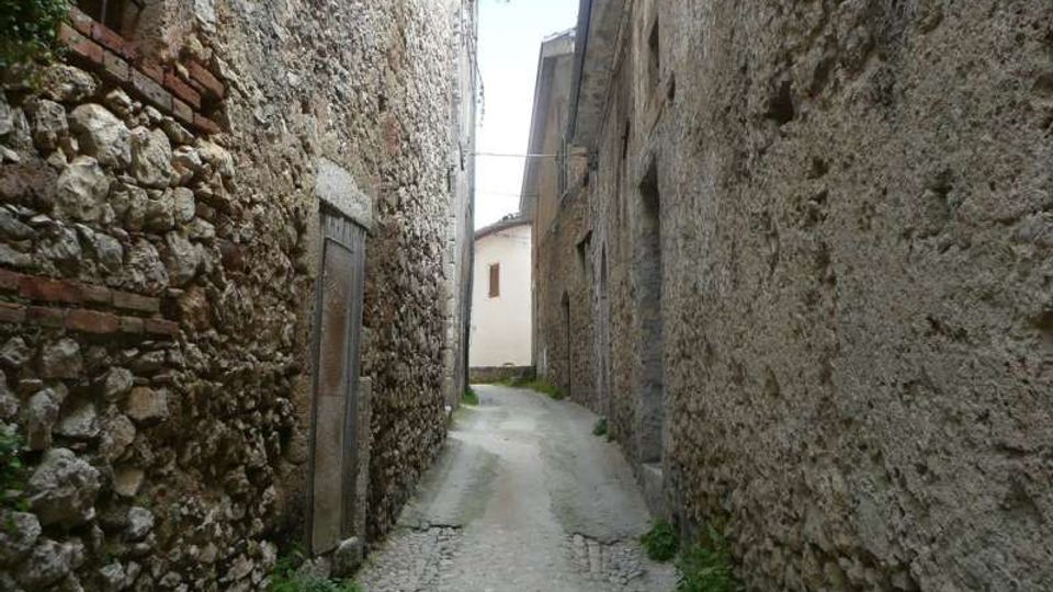 Vicolo stretto con pavimentazione in pietra tra alti edifici in muratura grezza e pietra, con porte e piccole finestre sulle facciate. Il passaggio curva leggermente e conduce verso una casa chiara in fondo, con vegetazione che cresce ai bordi.