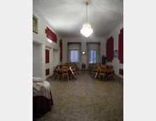 Sala interna ampia con pavimento in terrazzo, pareti bianche con pannelli rossi e un grande lampadario centrale. Ai lati sono impilate sedie in legno e sul fondo ci sono due finestre con tendine e una piccola applique a parete.