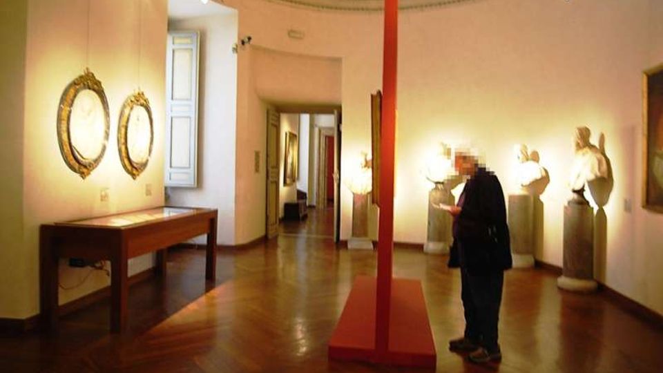 Interno di una galleria museale con pareti bianche e pavimento in parquet, illuminata da faretti a soffitto. Lungo le pareti sono esposti specchi con cornici dorate, una teca su tavolo e sculture su piedistalli, con un elemento espositivo rosso al centro della sala e un passaggio verso altre stanze sul fondo.
