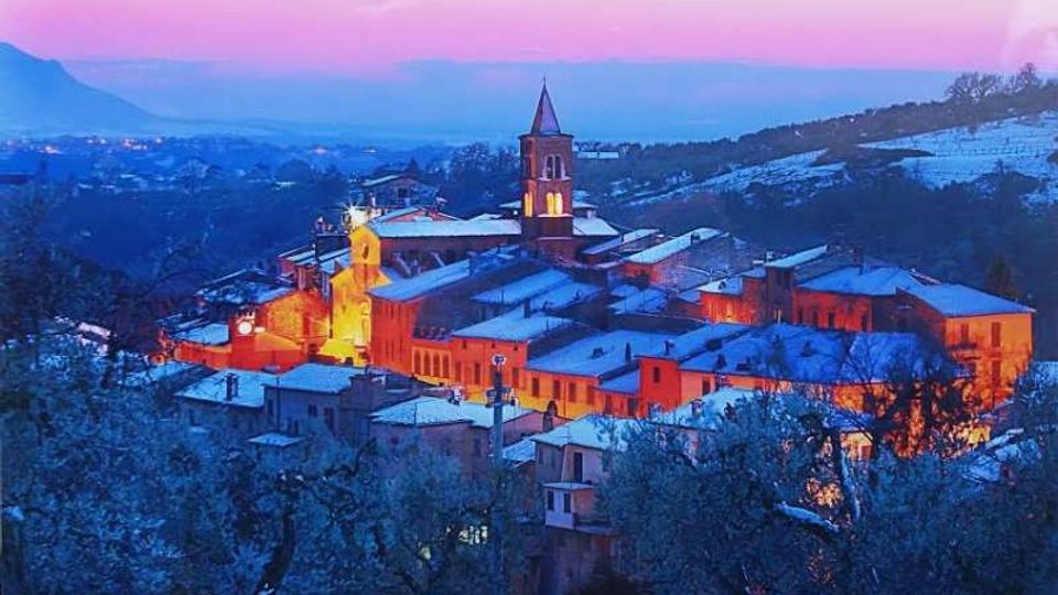 Veduta panoramica di un borgo collinare al crepuscolo, con tetti coperti di neve e un campanile illuminato al centro del paese. Sullo sfondo si vedono colline e montagne in lontananza sotto un cielo rosa e azzurro.