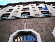 Facciata di un edificio istituzionale in pietra vista dal basso, con più piani di finestre rettangolari e un grande portale ad arco con inferriate. Due aste portabandiera sulla parete espongono bandiere, e una scritta incisa corre lungo la fascia sopra l’ingresso.