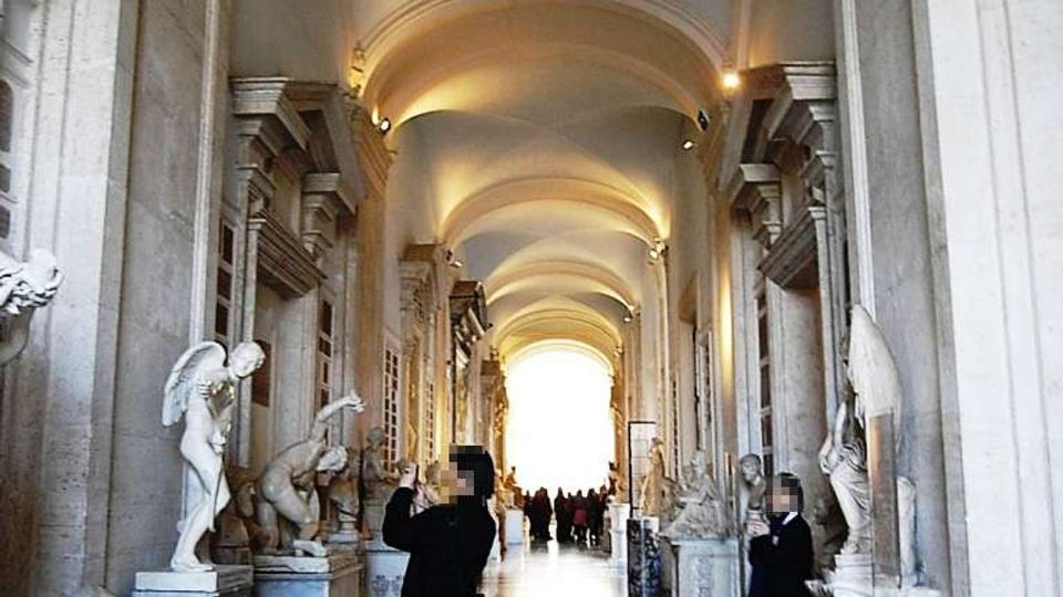 Lungo corridoio monumentale in pietra con alte arcate e soffitto a volte illuminato da luci calde. Ai lati si trovano nicchie e piedistalli con statue classiche in marmo e pavimento a motivi geometrici che conduce verso un’uscita luminosa in fondo.