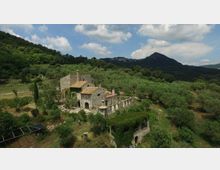 Veduta aerea di un casale in pietra con tetti in tegole e un’area porticata ad archi, circondato da uliveti e vegetazione collinare. Sullo sfondo si vedono montagne boscose sotto un cielo parzialmente nuvoloso.