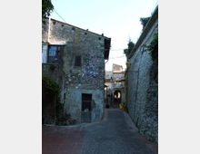 Vicolo stretto in un borgo storico, con pavimentazione in pietra e alti muri di case in muratura grezza. In fondo si vede un arco che attraversa la strada, mentre su una facciata a sinistra sono presenti decorazioni a mosaico e piatti ceramici.